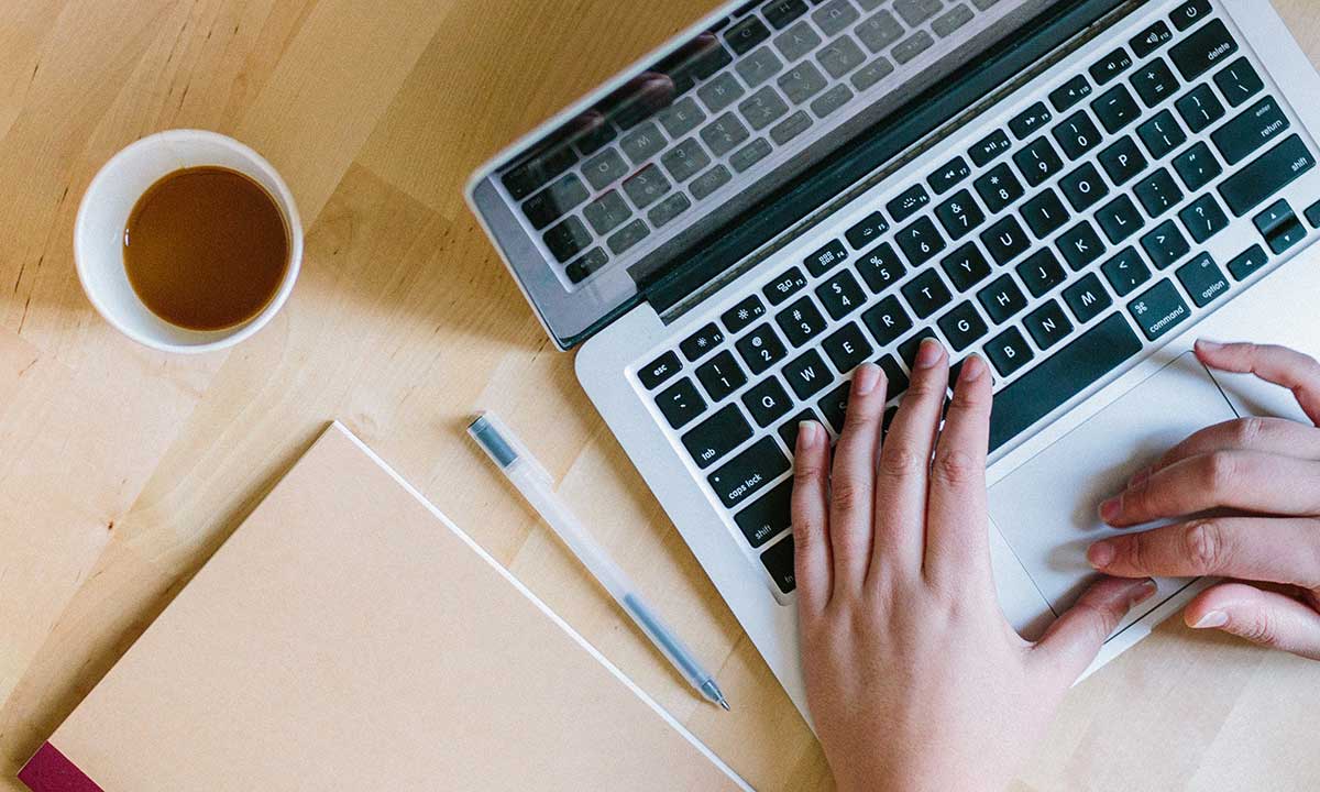 Händer skriver på en dator med anteckningsblock, penna och kaffemugg på bordet.